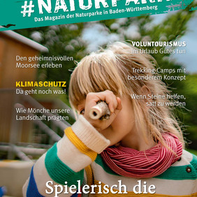 Titelbild des #Naturpark Magazins. Darauf ist ein Kind in einem bunten Wollpullover abgebildet, dass durch einen hohlen Ast in die Kamera schaut.