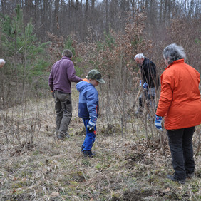 Foto von mehreren Personen, die auf einer Wiesenfläche stehen. Im Hintergrund steht unbelaubter Wald mit ein paar grünen Nadelbäumen dazwischen. Fast alle der sechs Personen sind von hinten zu sehen, rechts hat jemand einen Spaten in der Hand, links jemand eine Heckenschere. Alle haben Handschuhe an.