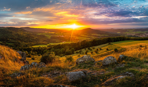 Foto eines Sonnenuntergangs, fotografiert von einem Hügel aus ins Tal. Im Hintergrund ist die tief stehende, orange leuchtende Sonne zu sehen. Davor ist eine weitläufige Hügellandschaft, die teilweise bewaldet ist. Es sind in der Ferne einige Siedlungen zu sehen. Im Vordergrund sind Felsen, die von hohen Gräßern umgeben sind.