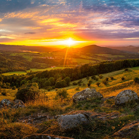 © VDN-Fotoportal/Jens Becker Foto eines Sonnenuntergangs, fotografiert von einem Hügel aus ins Tal. Im Hintergrund ist die tief stehende, orange leuchtende Sonne zu sehen. Davor ist eine weitläufige Hügellandschaft, die teilweise bewaldet ist. Es sind in der Ferne einige Siedlungen zu sehen. Im Vordergrund sind Felsen, die von hohen Gräßern umgeben sind.