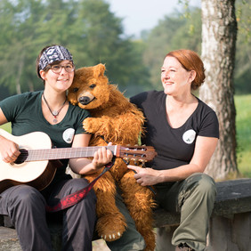 Kindertheater mit Brummelbutz, dem letzten Bär im Schönbuch. Foto: Figurentheater Martinshof Zwei Frauen mit Gitarre und Bär sitzen auf einer Bank